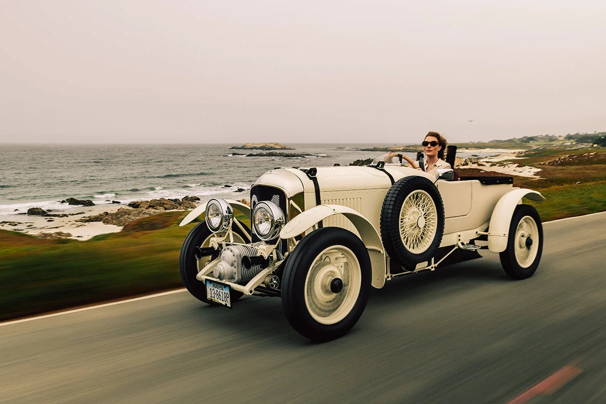 Cream-colored vintage open-top roadster traveling along a coastal road with the ocean and rocky shoreline behind it