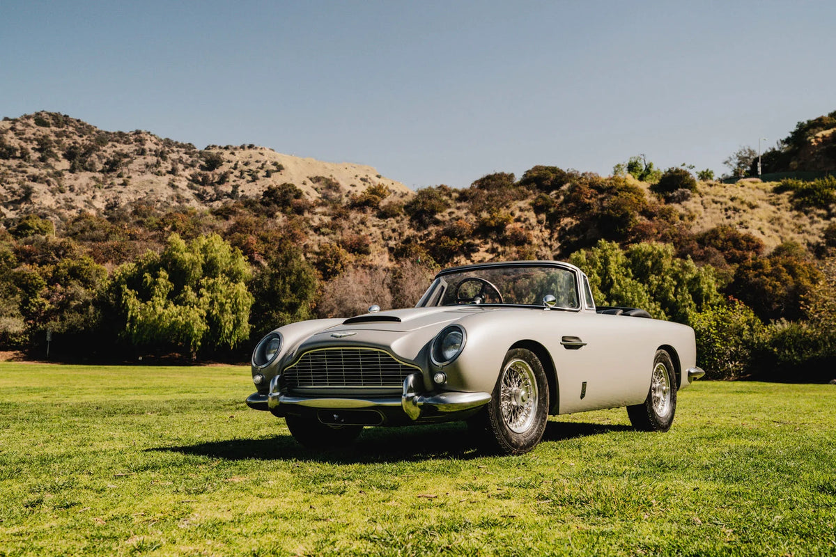 Classic silver convertible sports car parked on a grassy field beneath a clear blue sky.